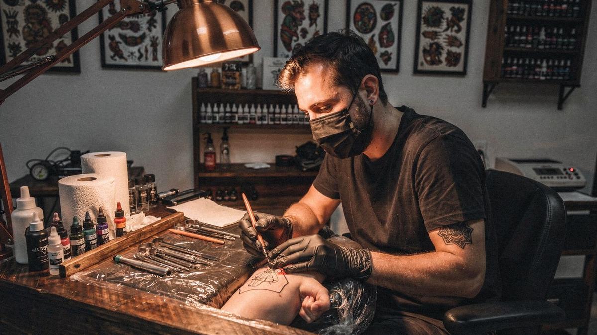 Artist working at a covered tattoo station at aspirelogically.com under a copper lamp, wearing gloves and a mask.